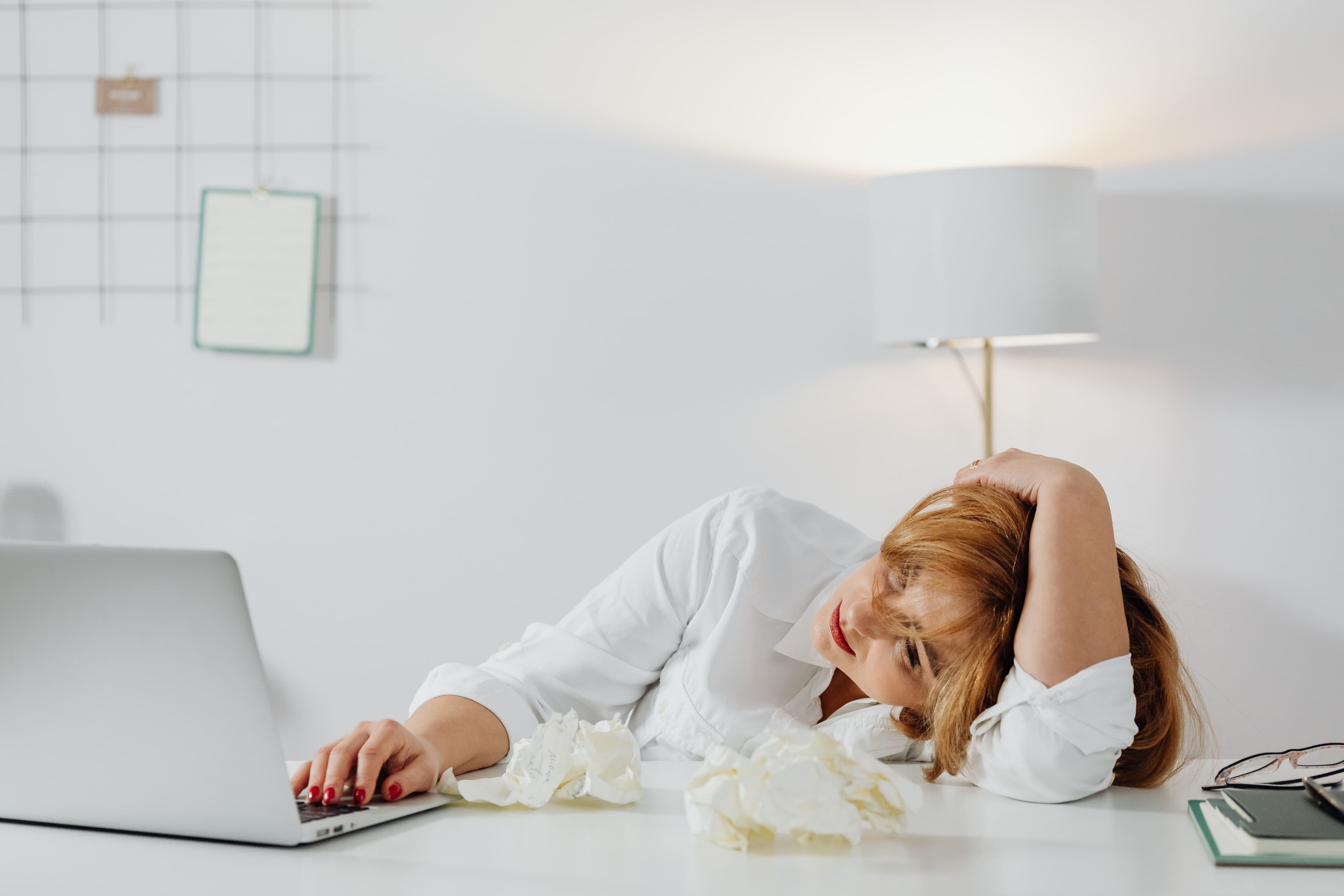 A woman lies on a desk in frustration or exhaustion next to a laptop